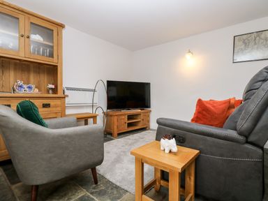 A living room with a grey armchair and recliner with red and green cushions a wooden cabinet wooden side tables and a television at Keepers Cottage in Liskeard