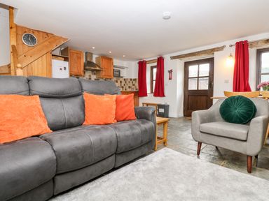 A living room with a gray sofa and orange cushions next to a kitchen with wooden cabinets at Keepers Cottage in Liskeard