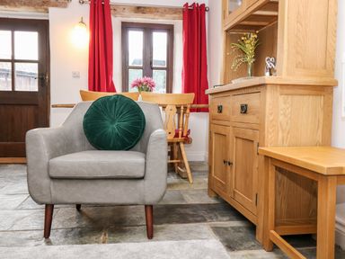 A room with a gray armchair with a green cushion a wooden cabinet a small wooden bench and a table with chairs near windows with red curtains at Keepers Cottage in Liskeard