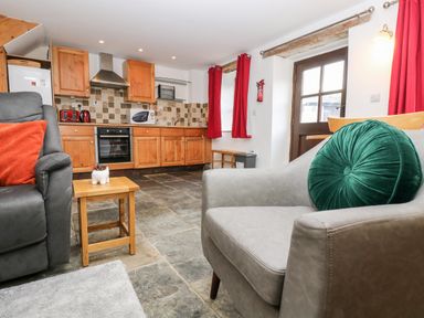 A kitchen with wooden cabinets and tiled backsplash next to a living area with gray chairs and red curtains at Keepers Cottage in Liskeard