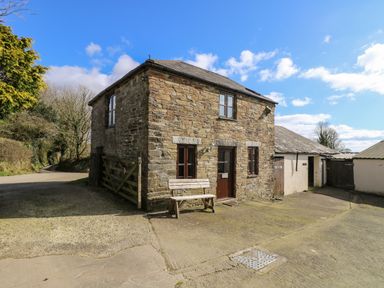 A stone cottage with wooden bench outside near a gated driveway and outbuildings under a blue sky at Keepers Cottage in Liskeard