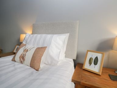 A bed with white pillows and a cushion with a deer design alongside wooden bedside tables with framed leaf pictures and lamps at Raybarrow in Windermere