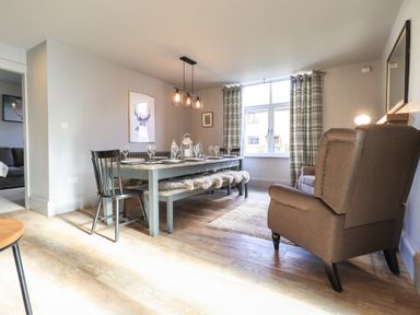 A dining room with a table set for six people with a bench and chairs a brown armchair near a window with plaid curtains at Raybarrow in Windermere