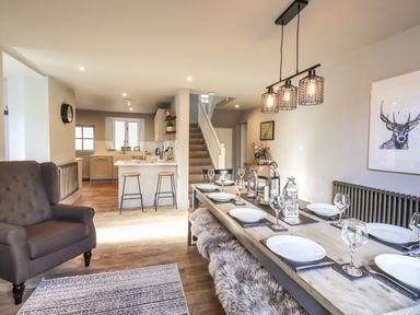 A dining table set with plates and glasses next to an armchair and open kitchen at Raybarrow in Windermere