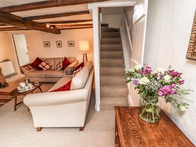 A living room with two sofas, a coffee table with tea set, a standing lamp, framed pictures on the wall, stairs, and a vase of flowers on a wooden table at Lavender Cottage in South Zeal