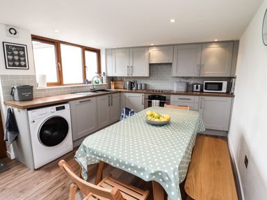 A kitchen with gray cabinets a washing machine a table with a polka dot tablecloth a bowl of fruit and wooden chairs at Lavender Cottage in South Zeal