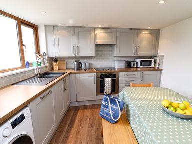 A kitchen with grey cabinets a wooden countertop a sink and washing machine a dining table with a polka dot tablecloth and a bowl of fruit at Lavender Cottage in South Zeal