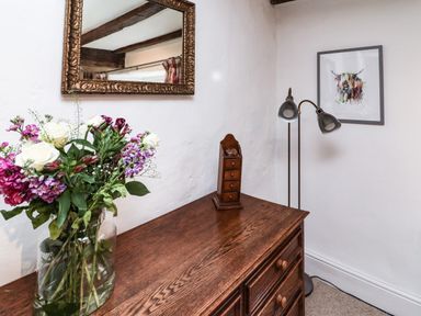 A wooden dresser with a vase of flowers and a small wooden chest under a mirror and next to a floor lamp and framed artwork at Lavender Cottage in South Zeal