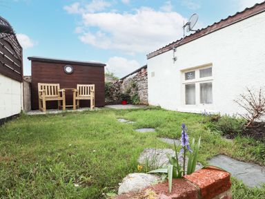 A small garden with grass, two wooden chairs and a table, a brown shed with a clock on it and a white building at Lavender Cottage in South Zeal