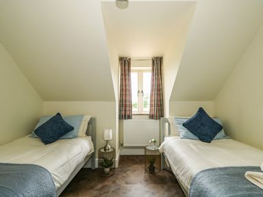 A bedroom with two single beds a window with plaid curtains and small tables with lamps and plants at The Attic in Blackborough End near King's Lynn