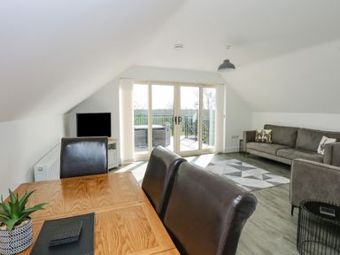 A dining table with chairs a gray sofa a small table and a TV in a living room with glass doors opening to a balcony at The Attic in Blackborough End near Kings Lynn