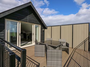 A balcony with a glass table and four wicker chairs outside a building with sliding glass doors and black siding at The Attic in Blackborough End near Kings Lynn