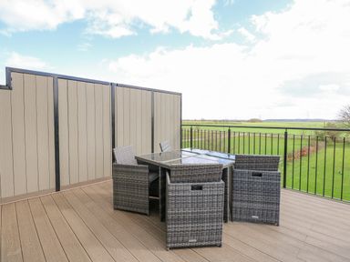 An outdoor patio with a glass table and four wicker chairs with a railing overlooking green fields at The Attic in Blackborough End near King's Lynn
