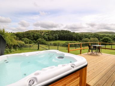 A deck with a hot tub and a table with chairs overlooking a grassy field and trees at Hwyl Rhayader near Llandrindod Wells