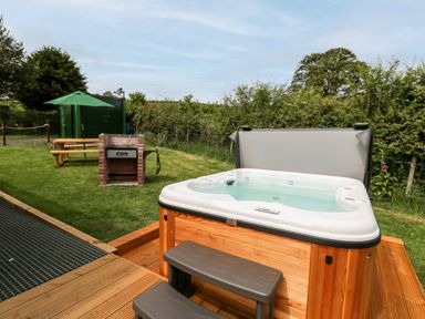 An outdoor area with a hot tub wooden steps a brick barbecue picnic table with an umbrella and green shed at Hwyl Rhayader near Llandrindod Wells