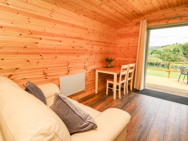 A wooden room with a cream sofa with cushions a small table with two chairs and a large window showing a green outdoor area at Hwyl Rhayader near Llandrindod Wells