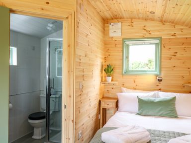 A bedroom with wooden walls a bed with white and green bedding a small wooden nightstand and a bathroom with a shower visible through an open door at Hwyl Rhayader near Llandrindod Wells