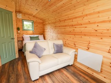 A small cabin room with wooden walls featuring a white sofa with cushions a bed with pillows and a green door at Hwyl Rhayader near Llandrindod Wells