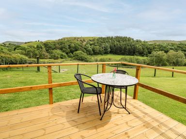 A wooden deck with a round mosaic table and two black chairs overlooking green fields and hills at Hwyl in Rhayader near Llandrindod Wells