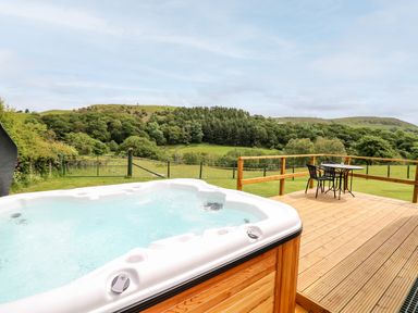 A hot tub on a wooden deck with a table and two chairs overlooking green fields and hills at Hwyl Rhayader near Llandrindod Wells