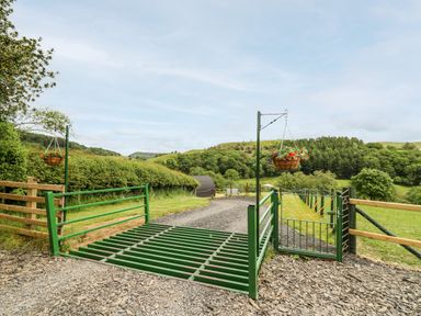 A green metal cattle grid and gate on a gravel path with hanging flower baskets and hills in the background at Hwyl in Rhayader near Llandrindod Wells