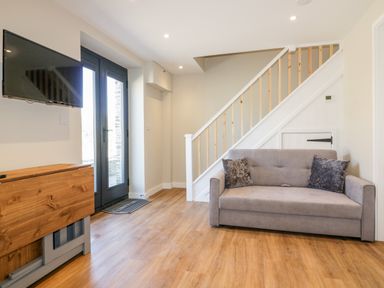 A living room with a grey sofa two cushions wooden floor a foldable wooden table stairs and a wall mounted TV at The Cornstore in Helland near Bodmin