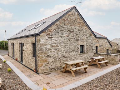 An exterior view of a stone building with a sloped roof and two wooden picnic tables on a paved patio at The Cornstore in Helland near Bodmin