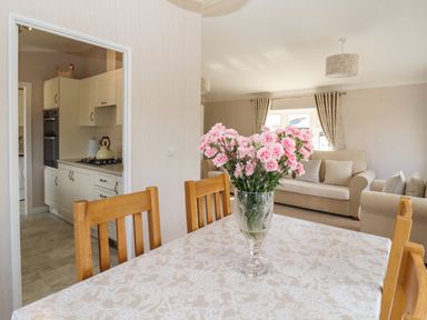 A dining area with a table covered by a patterned cloth and a glass vase with pink flowers and a kitchen visible through an open doorway at 12 Bucklesham Park in Bucklesham near Trimley St Martin