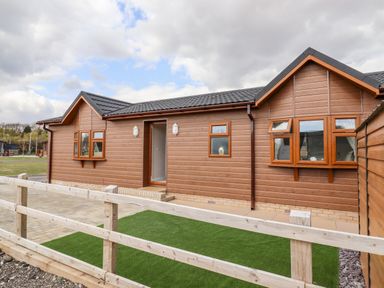 An exterior view of a brown wooden house with multiple windows a door and a small fenced area with artificial grass at 12 Bucklesham Park in Bucklesham near Trimley St Martin