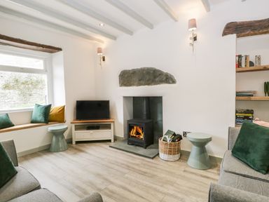 A living room with a wood burning stove two sofas a TV on a stand window seat with cushions and built in shelves at Turn Cottage in Satterthwaite near Hawkshead