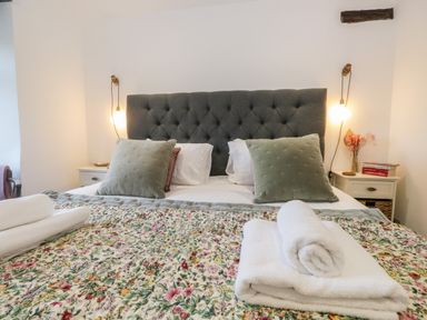 A bedroom with a grey tufted headboard two green pillows floral bedspread and rolled white towels at Turn Cottage in Satterthwaite near Hawkshead