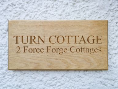 A wooden sign engraved with Turn Cottage 2 Force Forge Cottages on a textured white wall at Turn Cottage in Satterthwaite near Hawkshead