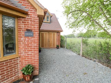 The exterior of a brick cottage with a wooden garage door a gravel driveway and a wire fence next to greenery at Globe Cottage in Stratford-Upon-Avon