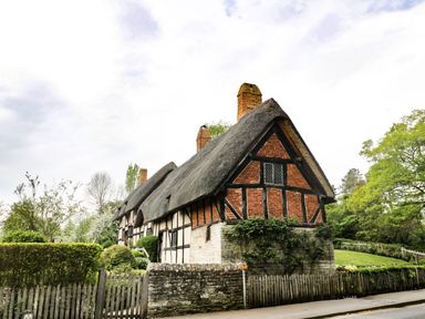 A traditional thatched roof cottage with timber framing surrounded by greenery and a wooden fence at Globe Cottage in Stratford-Upon-Avon