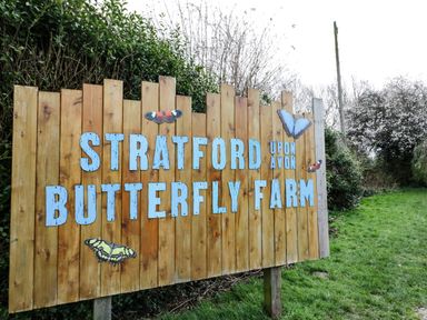 A wooden sign for Stratford upon Avon Butterfly Farm with butterflies on grass and trees in Stratford-Upon-Avon
