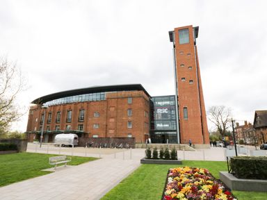 A large brick building with a tall tower and glass entrance next to a flower bed and green lawn at Globe Cottage in Stratford-Upon-Avon