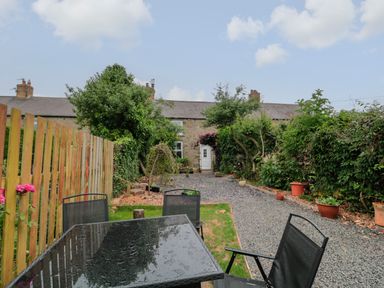 A garden with a gravel path green grass chairs and table in front of a stone house at Maple Tree Cottage in Longframlington