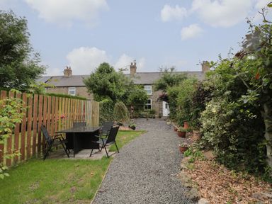 A gravel pathway leading to a stone house with garden chairs and table on the left at Maple Tree Cottage in Longframlington