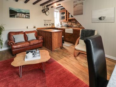 A living room with a brown leather sofa a wooden coffee table a wooden kitchen with cabinets and a sink and chairs at Maple Tree Cottage in Longframlington