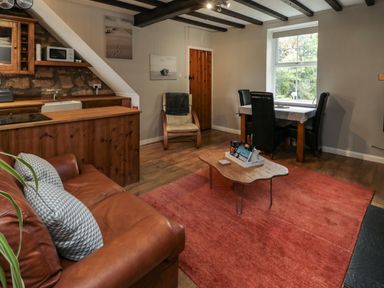 A living room with a brown leather sofa a wooden coffee table a dining table with black chairs and a wooden kitchen area at Maple Tree Cottage in Longframlington