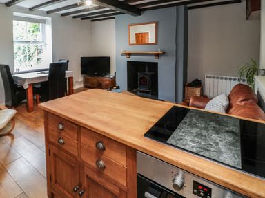 A kitchen island with stove top beside a living area with a fireplace, dining table, television and leather sofa at Maple Tree Cottage in Longframlington