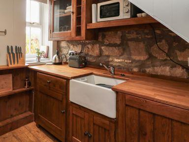 A kitchen with wooden cabinets a white sink a microwave a kettle a toaster and knives on a stand at Maple Tree Cottage in Longframlington