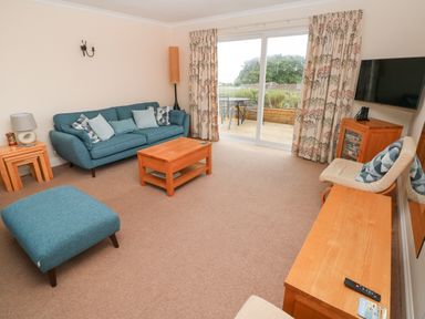 A living room with blue sofa chairs wooden tables and a TV by sliding glass doors at Trem Y Morfa in Newport Pembrokeshire