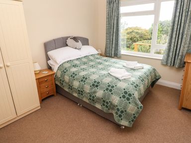 A bedroom with a bed covered by a patterned blanket two folded towels on the bed a wardrobe two wooden bedside tables with lamps curtains and a large window at Trem Y Morfa in Newport Pembrokeshire