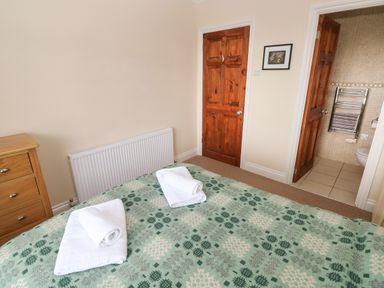 A bedroom with a bed covered by a patterned blanket and two rolled towels a wooden drawer unit and two wooden doors one leading to a tiled bathroom at Trem Y Morfa Newport Pembrokeshire