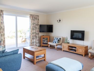 A living room with blue chairs wooden tables wall mounted televisions and sliding glass doors at Trem Y Morfa in Newport Pembrokeshire