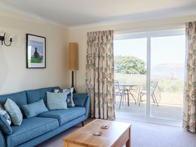 A living room with a blue sofa pillows wooden coffee table and sliding glass door leading to a patio with metal chairs at Trem Y Morfa in Newport Pembrokeshire