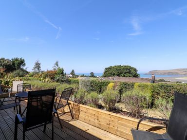 A wooden deck with black chairs and a table overlooking bushes houses and a distant coastline at Trem Y Morfa in Newport Pembrokeshire