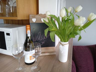 A kitchen counter with a microwave bottle of wine two wine glasses a vase of white tulips and a welcome sign at Silver Birch Lodge in Misterton