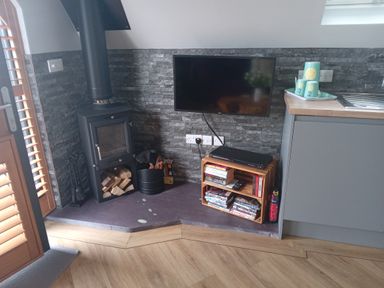 A corner with a wood burning stove firewood and tools a wall mounted TV and a small wooden shelf with books and DVDs at Silver Birch Lodge in Misterton